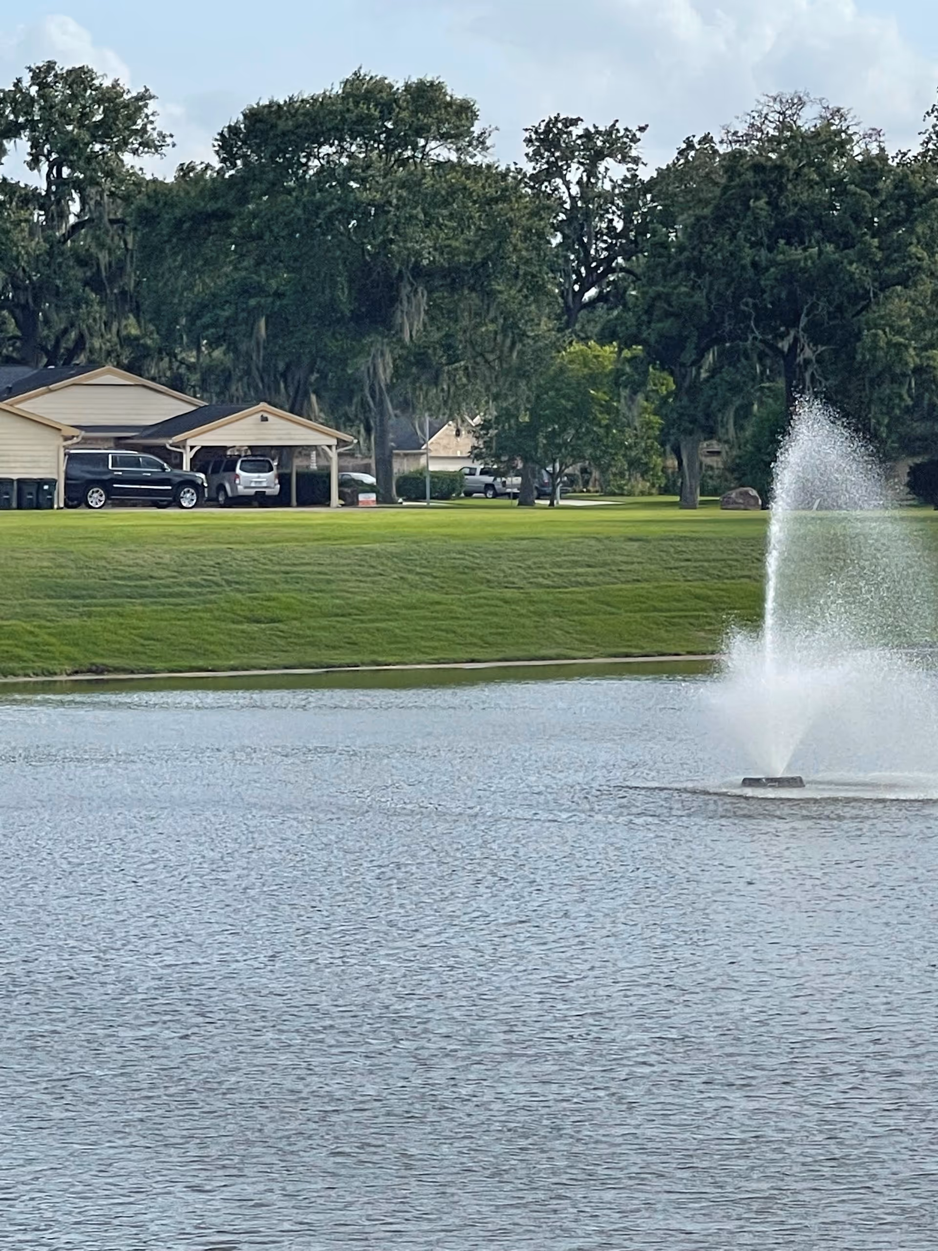 A serene outdoor scene featuring a pond with a water fountain spraying water upwards. In the background, there is a grassy area with several large trees and a few parked vehicles near a small building with a covered parking area.