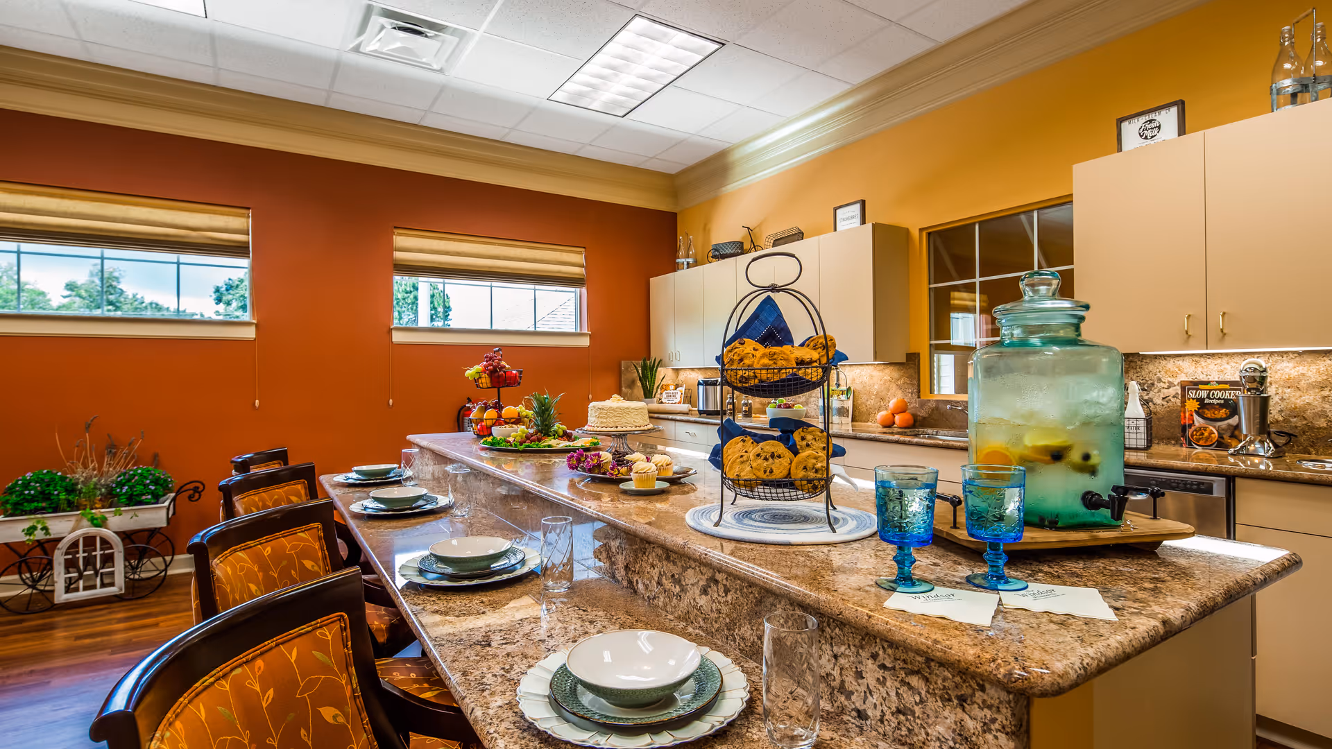 A bright kitchen area with a long marble countertop set with plates, bowls, glasses, and blue goblets. The countertop features a tiered tray of cookies, a large glass dispenser with infused water, and various plates of food. The kitchen has beige cabinets, a window, and warm orange walls with two small windows letting in natural light. There are several chairs with orange patterned cushions around the counter.