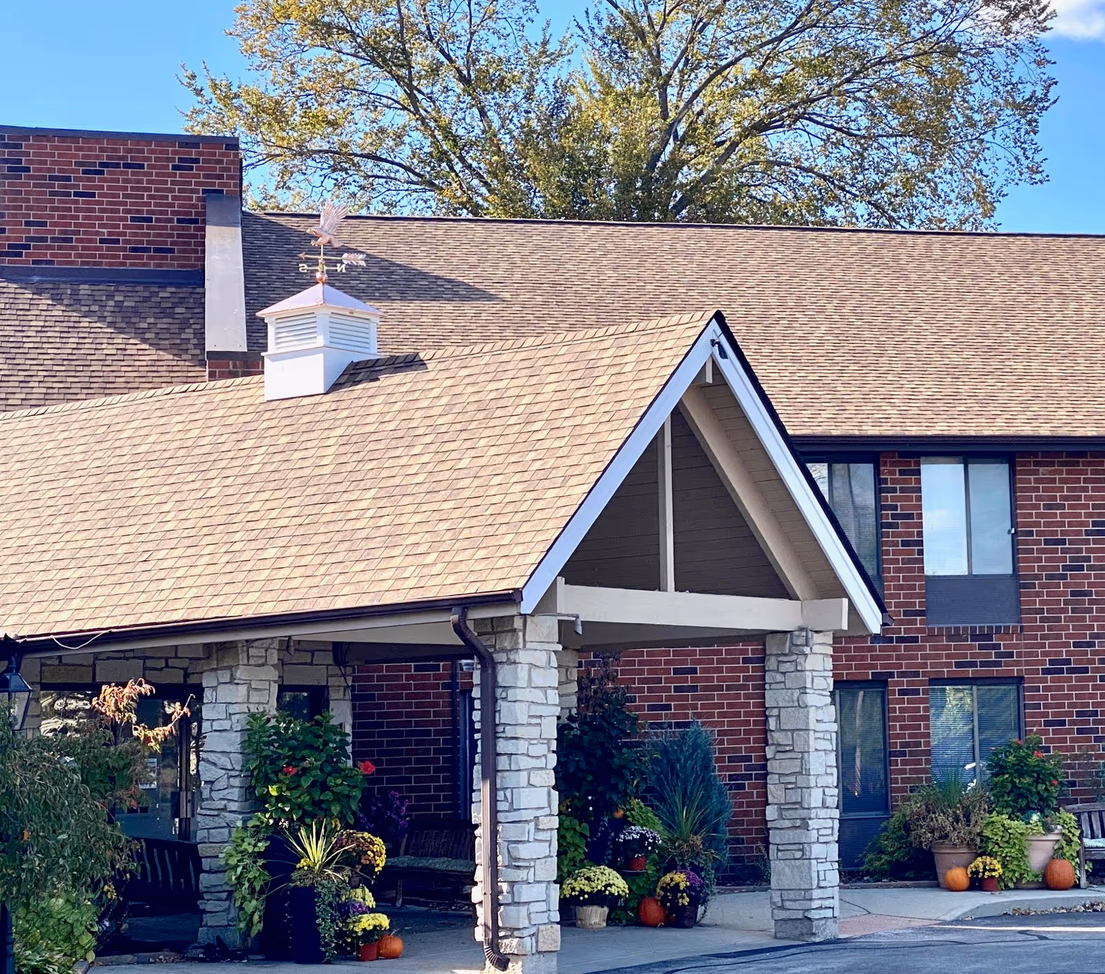 Entrance of a senior living facility named Independence Village featuring a covered driveway supported by stone pillars, with various potted plants and pumpkins decorating the area. The building has a brick exterior and a shingled roof with a weather vane on top. Trees and a clear blue sky are visible in the background.