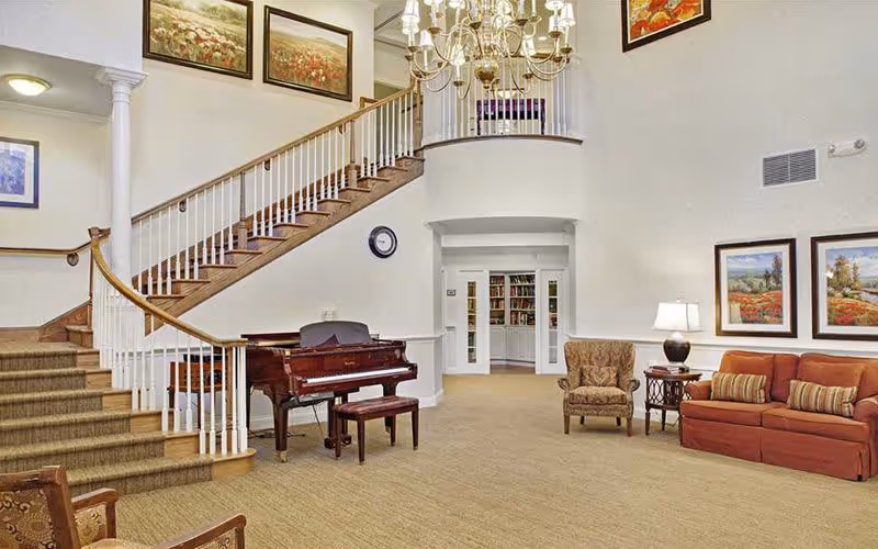 Spacious senior living facility common area with a grand staircase, a polished wooden piano with a matching bench, a comfortable armchair, a red sofa with striped cushions, a side table with a lamp, and several framed landscape paintings on the walls. A chandelier hangs from the ceiling, and a doorway leads to a room with bookshelves.
