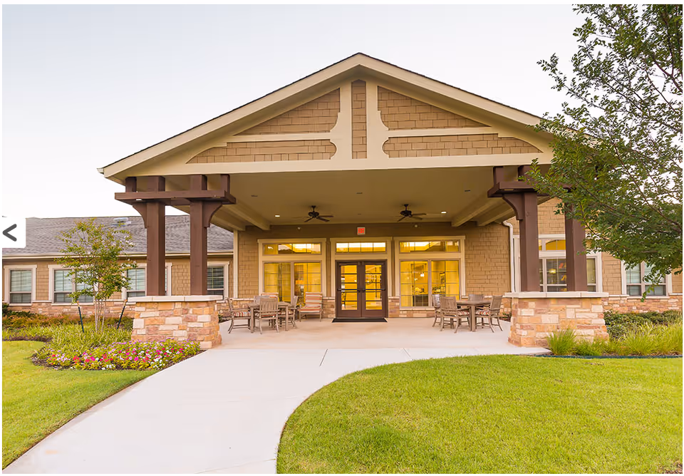 Front entrance of a senior living facility with a covered porch supported by wooden beams and stone pillars. There are outdoor tables and chairs on the porch, surrounded by well-maintained green grass and flower beds.