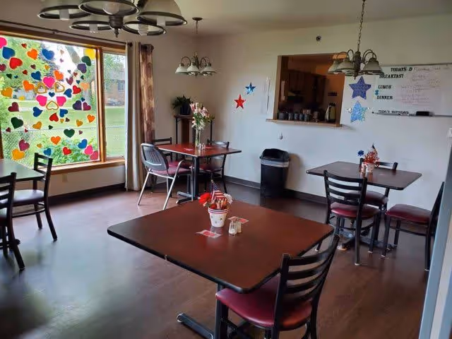 A dining room with several square tables and chairs arranged neatly. The room has a large window decorated with colorful heart-shaped stickers. There are light fixtures hanging from the ceiling, a trash bin near a pass-through window to the kitchen, and a whiteboard on the wall displaying meal information. The floor is wooden, and the room is well-lit with natural light.