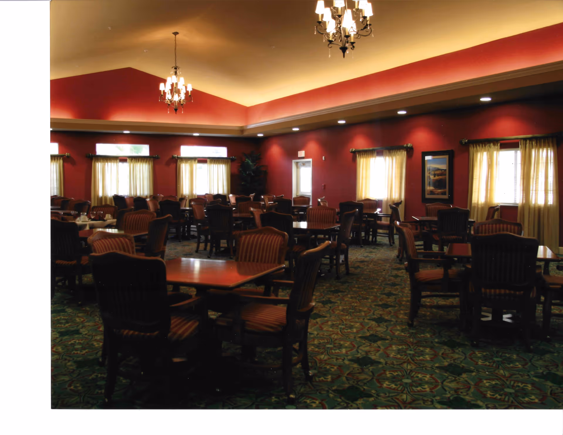 Spacious dining room with multiple wooden tables and upholstered chairs, red walls, chandeliers, and patterned carpet.