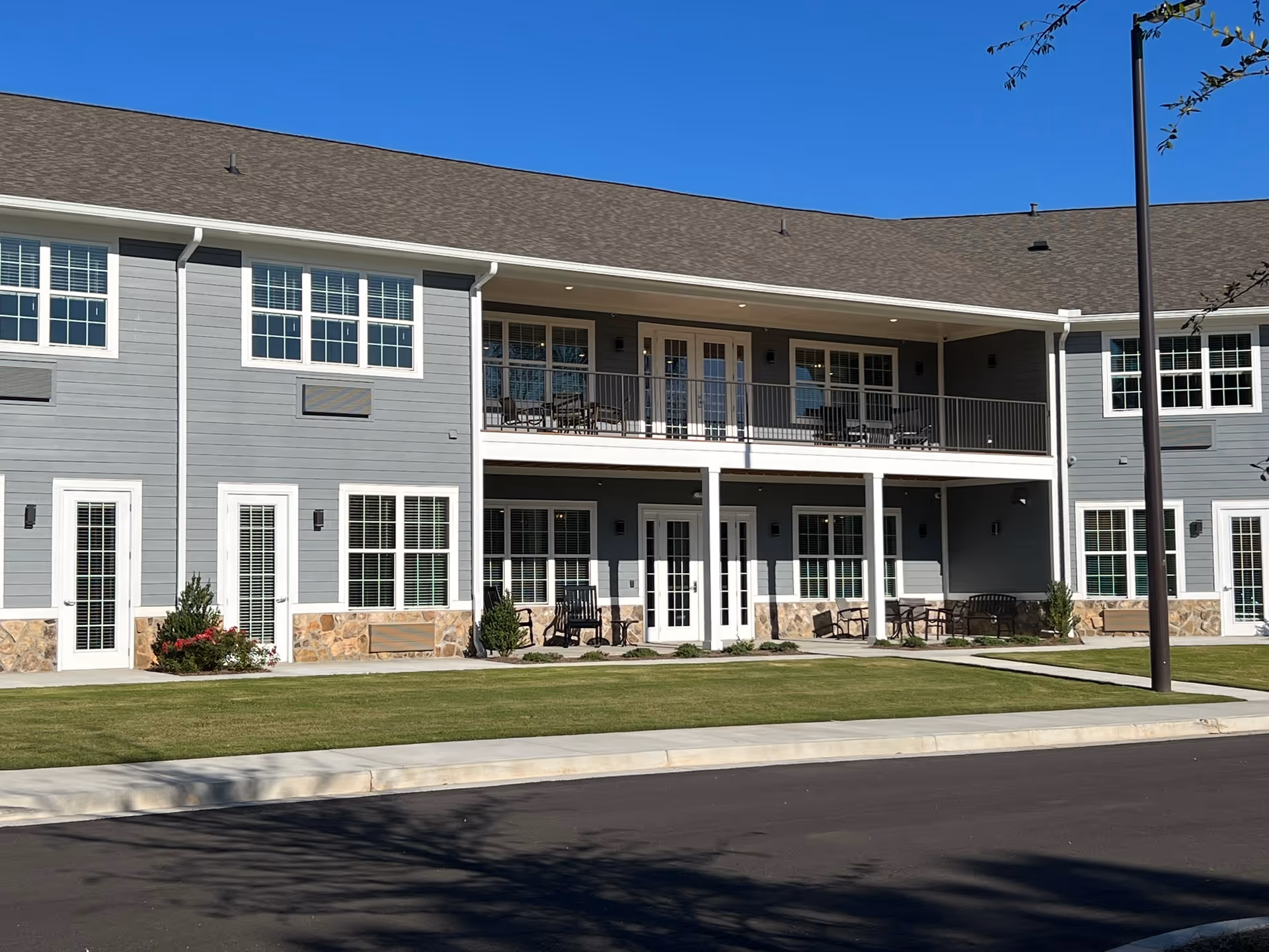 Exterior view of a two-story assisted living facility building with gray siding, white trim, and stone accents at the base. The building features multiple windows and doors, a covered balcony with outdoor seating on the second floor, and a covered patio area with benches and chairs on the ground floor. The foreground shows a well-maintained lawn and a paved driveway under a clear blue sky.