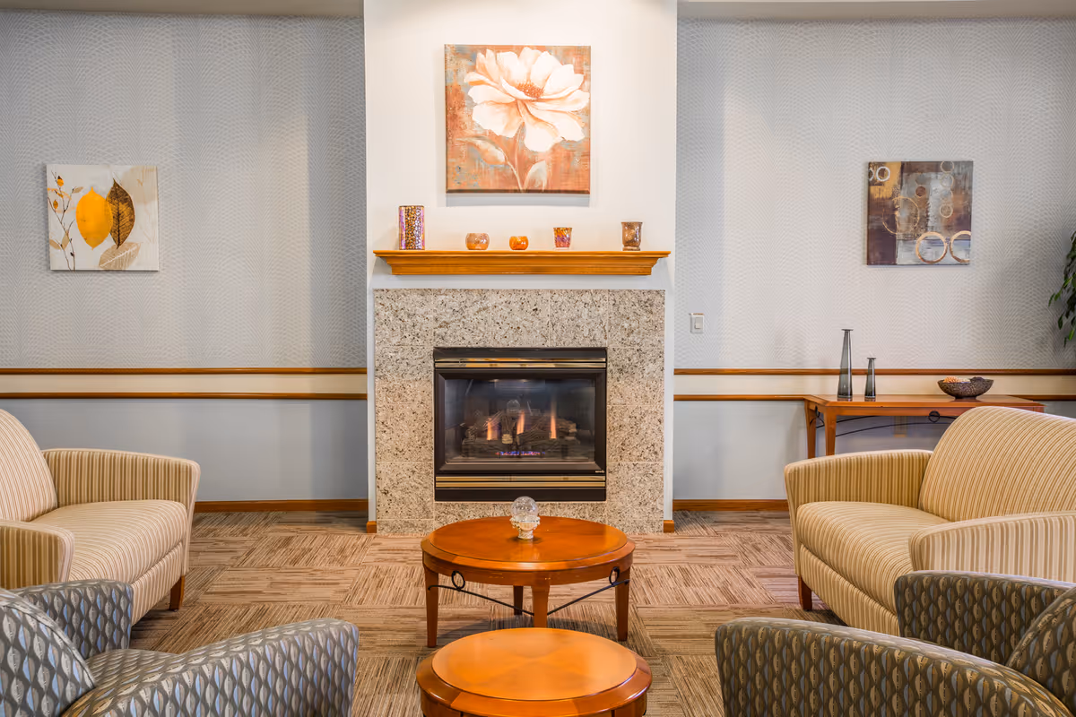 A cozy living room area with a lit fireplace in the center, flanked by two beige striped armchairs and two patterned armchairs. A round wooden coffee table and a smaller round wooden side table are in front of the fireplace. The walls have light-colored wallpaper with two framed artworks and a wooden console table with decorative items on the right side.