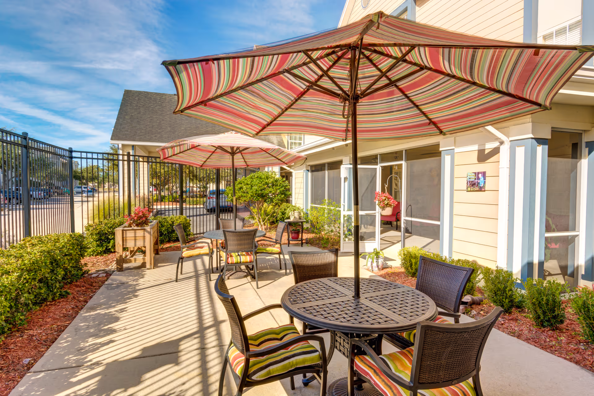 Sunlit outdoor patio with round tables, striped umbrellas, and chairs beside a fenced garden and building entrance.