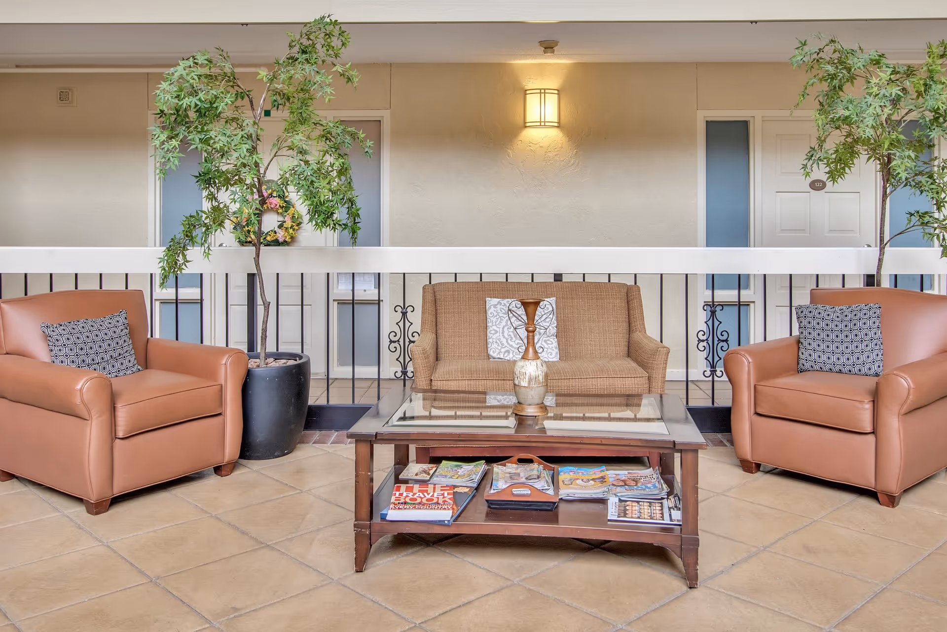 A cozy seating area in a hallway or common space with two brown leather armchairs, a brown fabric loveseat with two decorative pillows, a glass-top coffee table with magazines underneath, two potted plants, and a wall-mounted light fixture above. Doors to rooms are visible in the background.