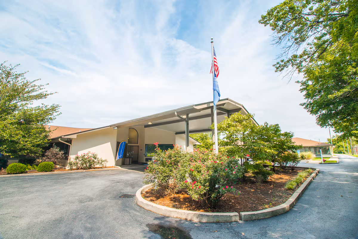 Entrance of a single-story senior living facility with a covered drop-off canopy, flagpole, and landscaped driveway.