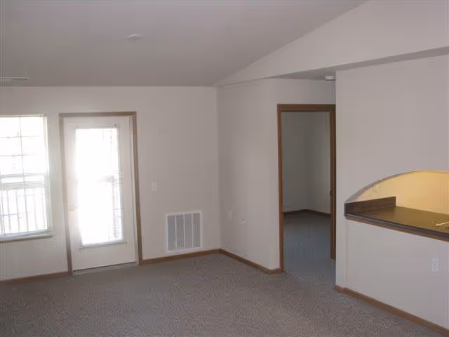 An empty interior room with beige walls and carpeted floor. There is a door with a window and blinds on the left side, a window next to it, and an open doorway leading to another room on the right. A partial view of a kitchen counter with a pass-through window is visible on the right side.