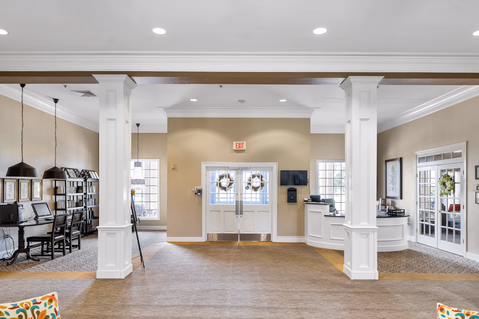 Interior view of a senior living facility lobby with beige walls, white pillars, and a carpeted floor. There is a reception desk on the right side, a set of double doors with wreaths in the center, and a seating or work area with chairs and hanging lamps on the left. The space is well-lit with recessed ceiling lights and natural light from windows.