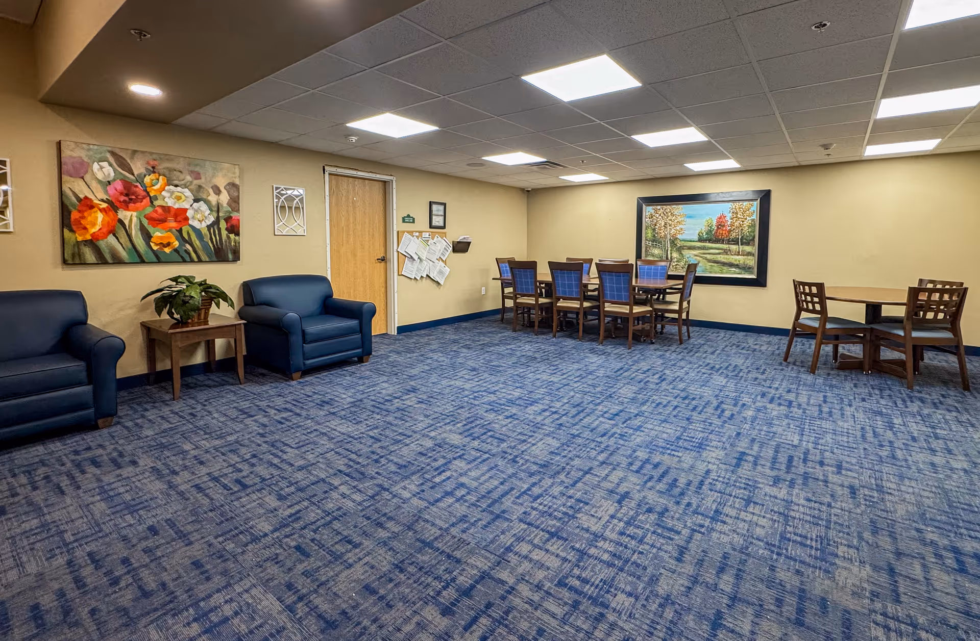 A spacious room with blue patterned carpet and beige walls featuring two blue armchairs with a small wooden table and plant between them on the left side. There are two sets of tables and chairs on the right side, one with six chairs around a rectangular table and another with four chairs around a round table. The walls are decorated with colorful paintings and a bulletin board near a wooden door.