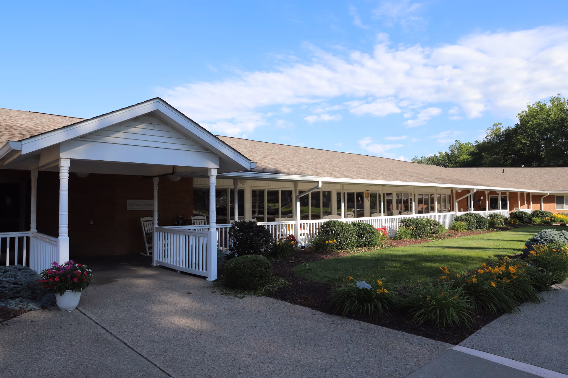 Exterior view of The Timbers of Jasper facility showing a long single-story building with a covered entrance, white railings, and a landscaped garden with bushes and flowers under a partly cloudy blue sky.