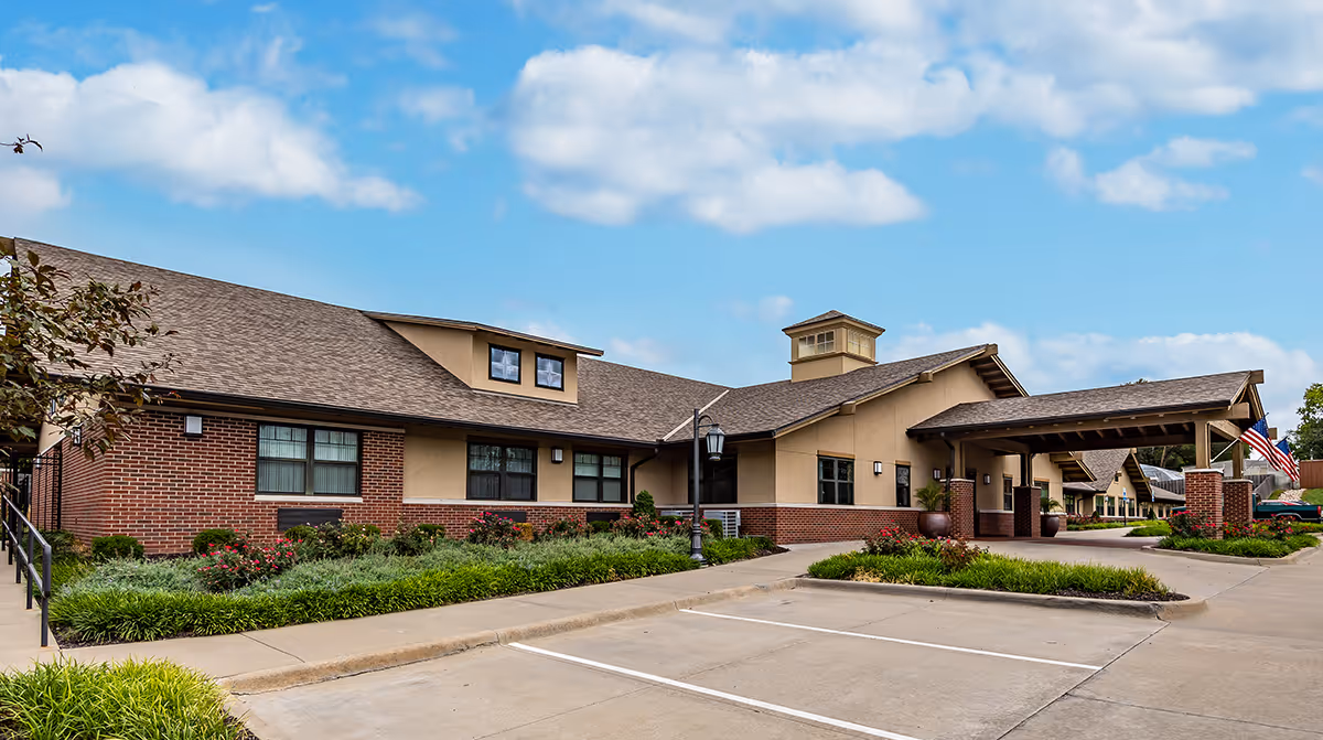 Exterior view of a single-story senior living facility building with a combination of brick and beige siding, a covered entrance, landscaped greenery, and a parking area under a partly cloudy sky.