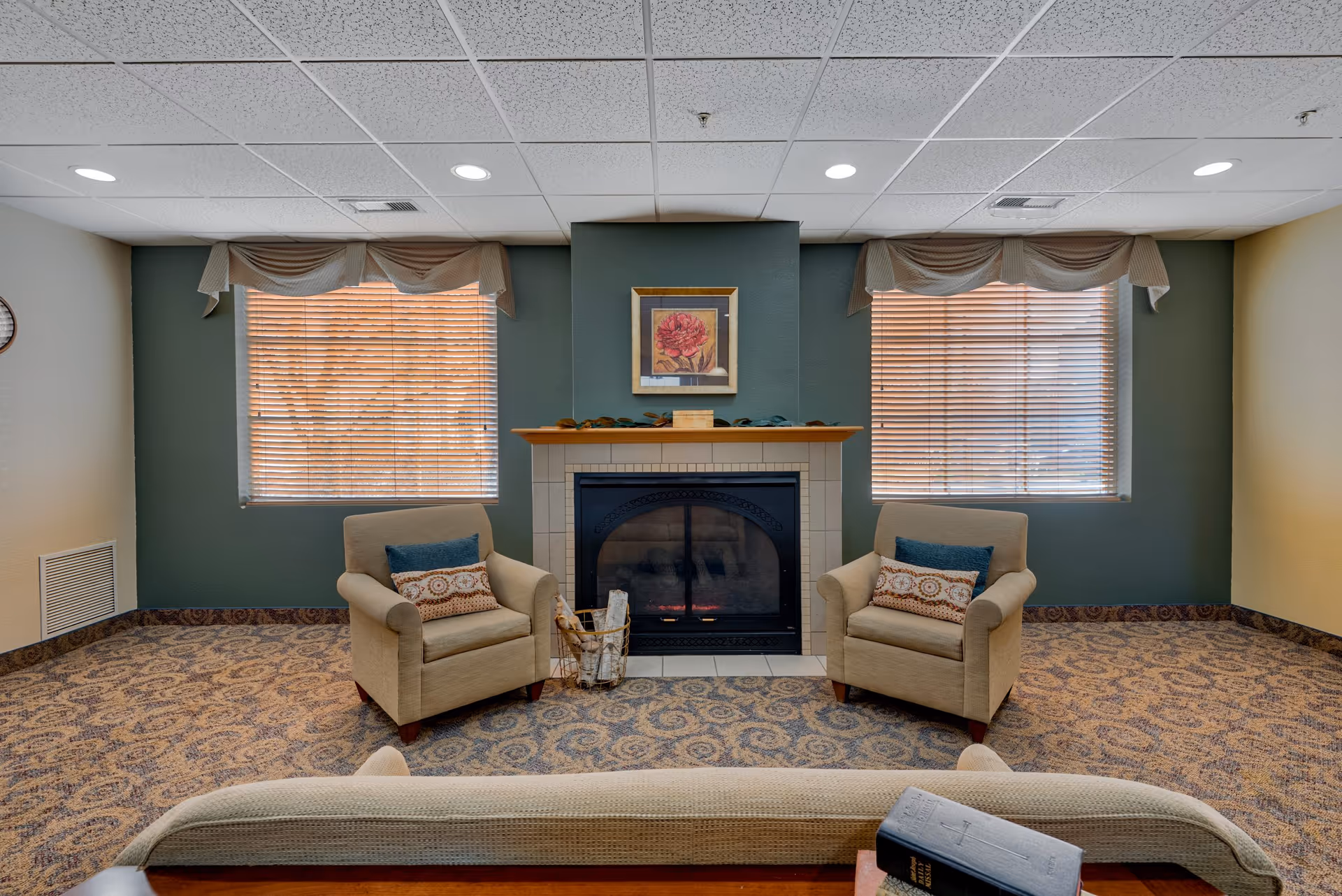 A cozy living room with two beige armchairs facing a fireplace with a wooden mantel. Above the fireplace is a framed picture of a red flower. Two windows with wooden blinds and beige valances flank the fireplace. The room has patterned carpet and a beige sofa partially visible in the foreground with a book resting on its arm.