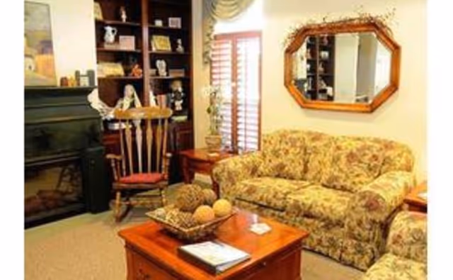 A cozy living room with floral patterned sofas, a wooden rocking chair, a wooden coffee table with decorative items, a bookshelf filled with various objects, a fireplace, and a wall mirror above one sofa. There is a window with wooden shutters and a side table with a plant.
