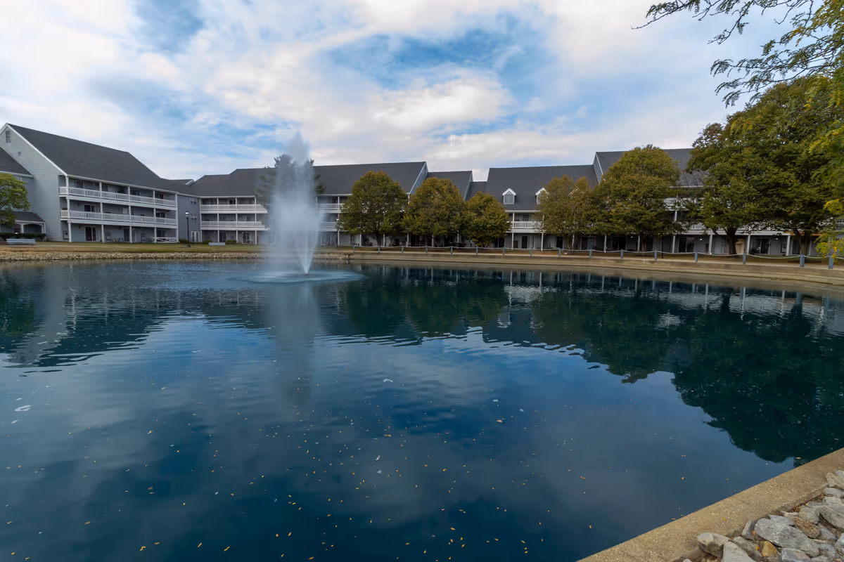 Three-story residential building with balconies facing a pond with a central fountain and trees.