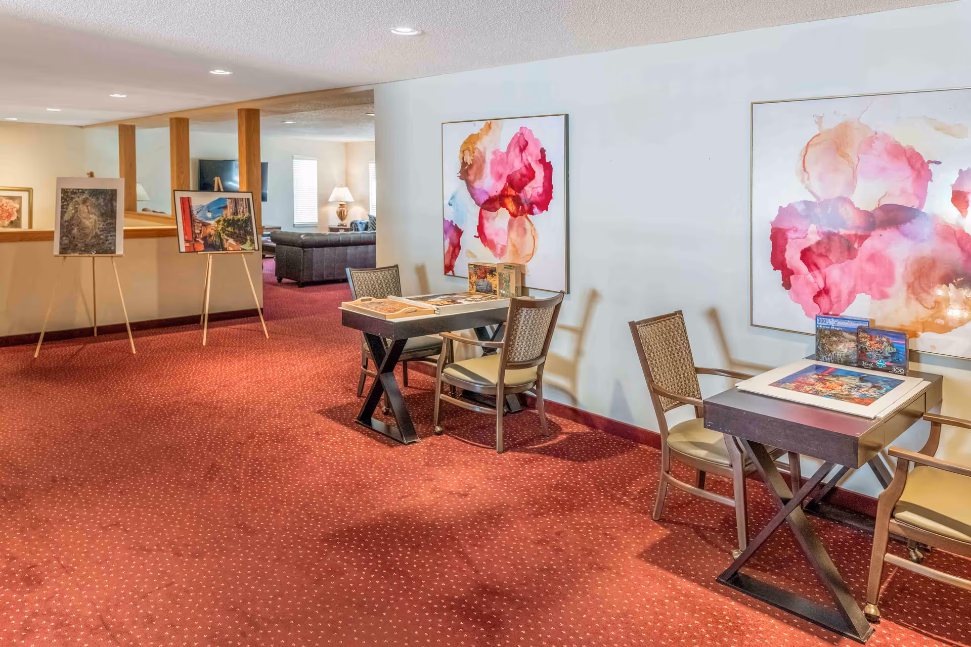 Carpeted communal interior with small tables and chairs for puzzles or games, colorful wall art, and a seating area in the background.