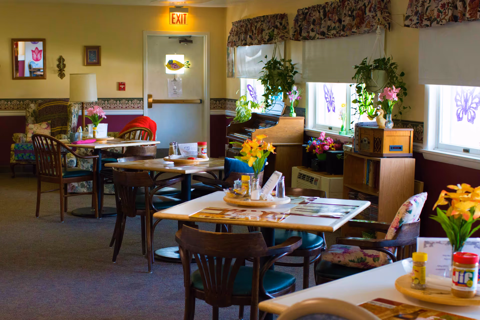 A cozy dining area in a senior living facility with several tables and chairs. The tables have flowers, condiments, and menus on them. There are large windows with floral valances and butterfly decorations, a piano with plants on top, and a bookshelf with more plants and flowers. An exit door is visible in the background.