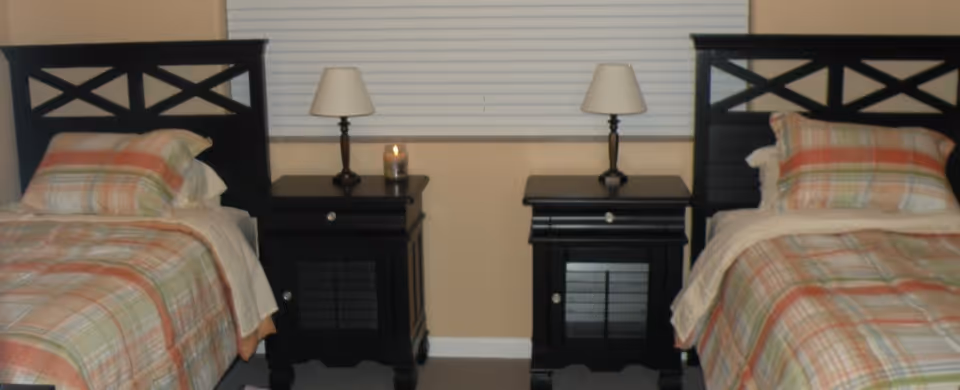 A bedroom with two twin beds featuring black wooden headboards and plaid bedding in shades of orange, green, and beige. Between the beds are two black nightstands, each with a small lamp. A candle is placed on the left nightstand. A window with closed white blinds is centered behind the nightstands.