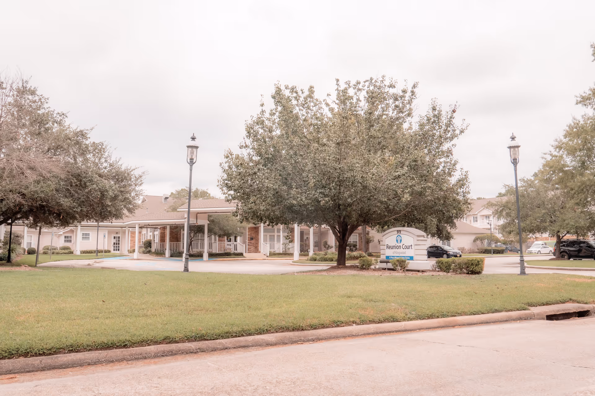 Exterior view of Reunion Court of Kingwood senior living facility showing a large tree, a sign with the facility name, lamp posts, a covered entrance, and parked cars in the background.