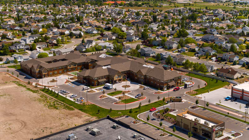 Aerial view of Summit Senior Living facility surrounded by a residential neighborhood. The building has a large parking lot with several cars parked, landscaped areas, and is situated near commercial buildings and open land.