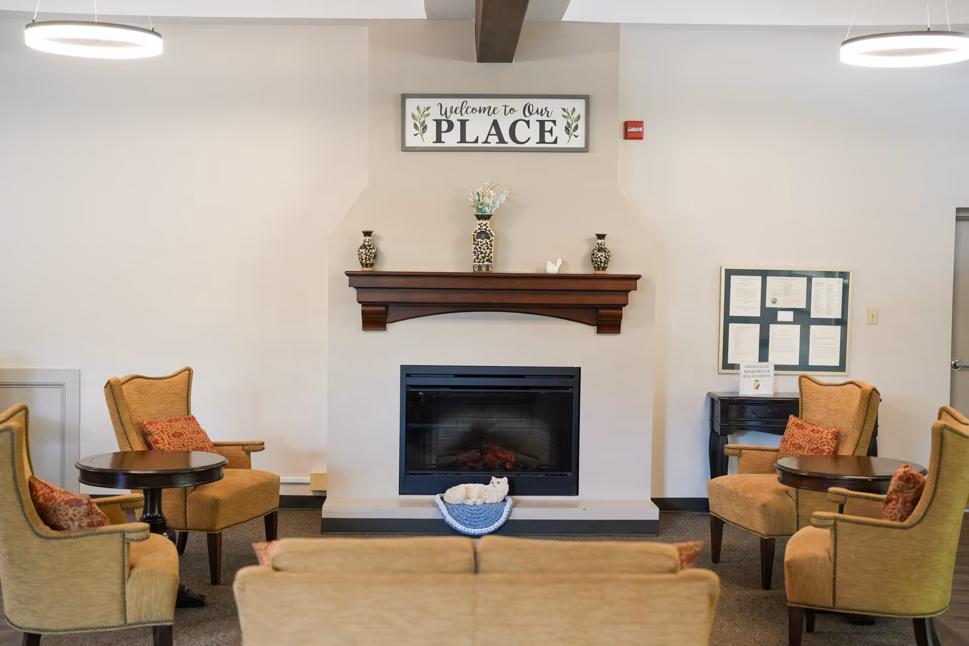 A cozy living room area with a fireplace in the center. Above the fireplace is a sign that reads 'Welcome to Our PLACE'. There are four upholstered armchairs with red patterned cushions arranged around two small round tables. A small decorative cat figure is placed on a blue mat in front of the fireplace. The walls are light-colored, and there is a bulletin board with papers on the right side.