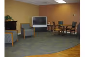 A common area in a senior living facility featuring a television on a stand against a beige wall, two upholstered armchairs with patterned fabric, a wooden table with four chairs, and a small table with a plant. The floor has a combination of carpet and hardwood.