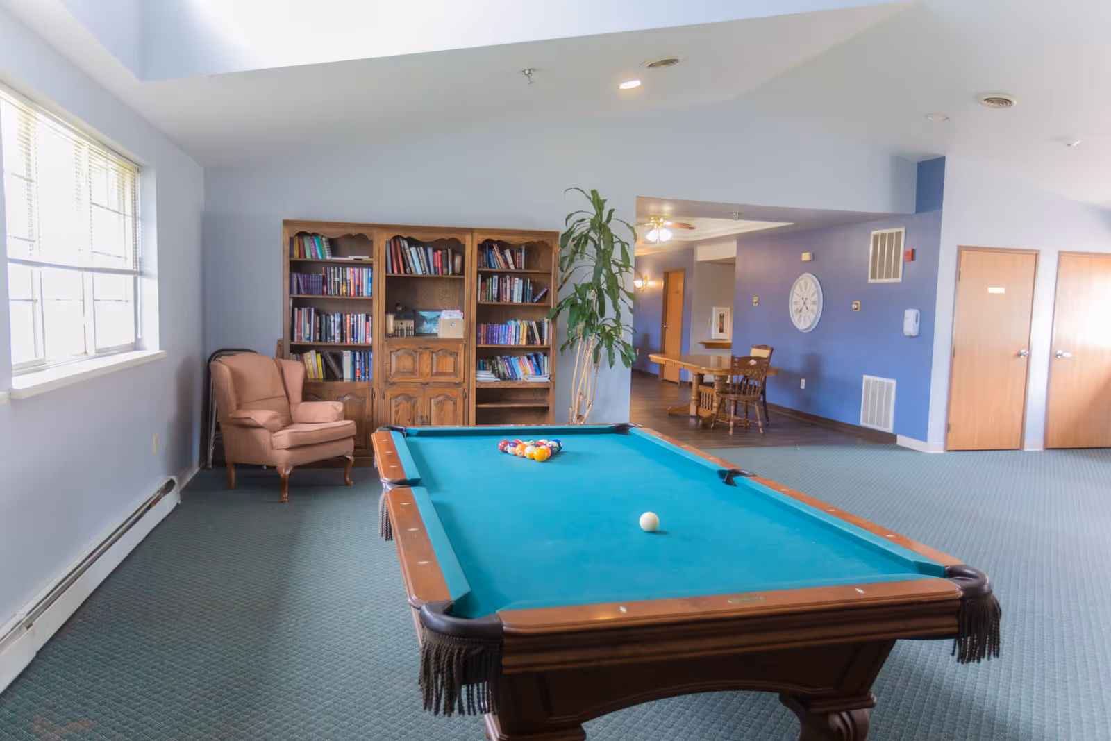 A recreational room in a senior living facility featuring a pool table with balls arranged for a game, a wooden bookshelf filled with books, a comfortable armchair, a large window letting in natural light, a tall indoor plant, and a dining area with a table and chairs in the background.