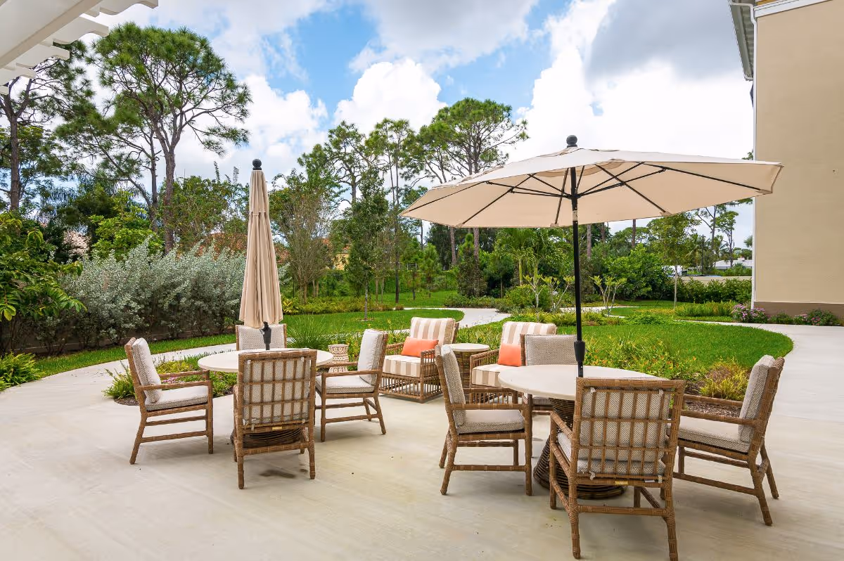 Outdoor patio with wicker chairs and tables under umbrellas on a paved area overlooking landscaped greenery and trees.
