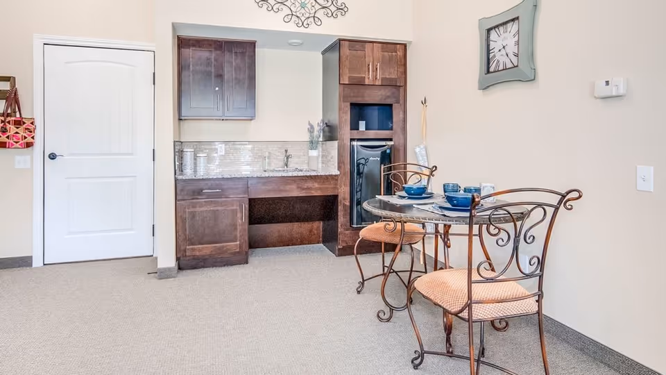 A small dining area in an assisted living facility featuring a round glass-top table with two wrought iron chairs with cushioned seats. Behind the table is a kitchenette with dark wood cabinets, a small refrigerator, a granite countertop, and a decorative backsplash. A wall clock and a decorative wall piece are visible on the beige walls.