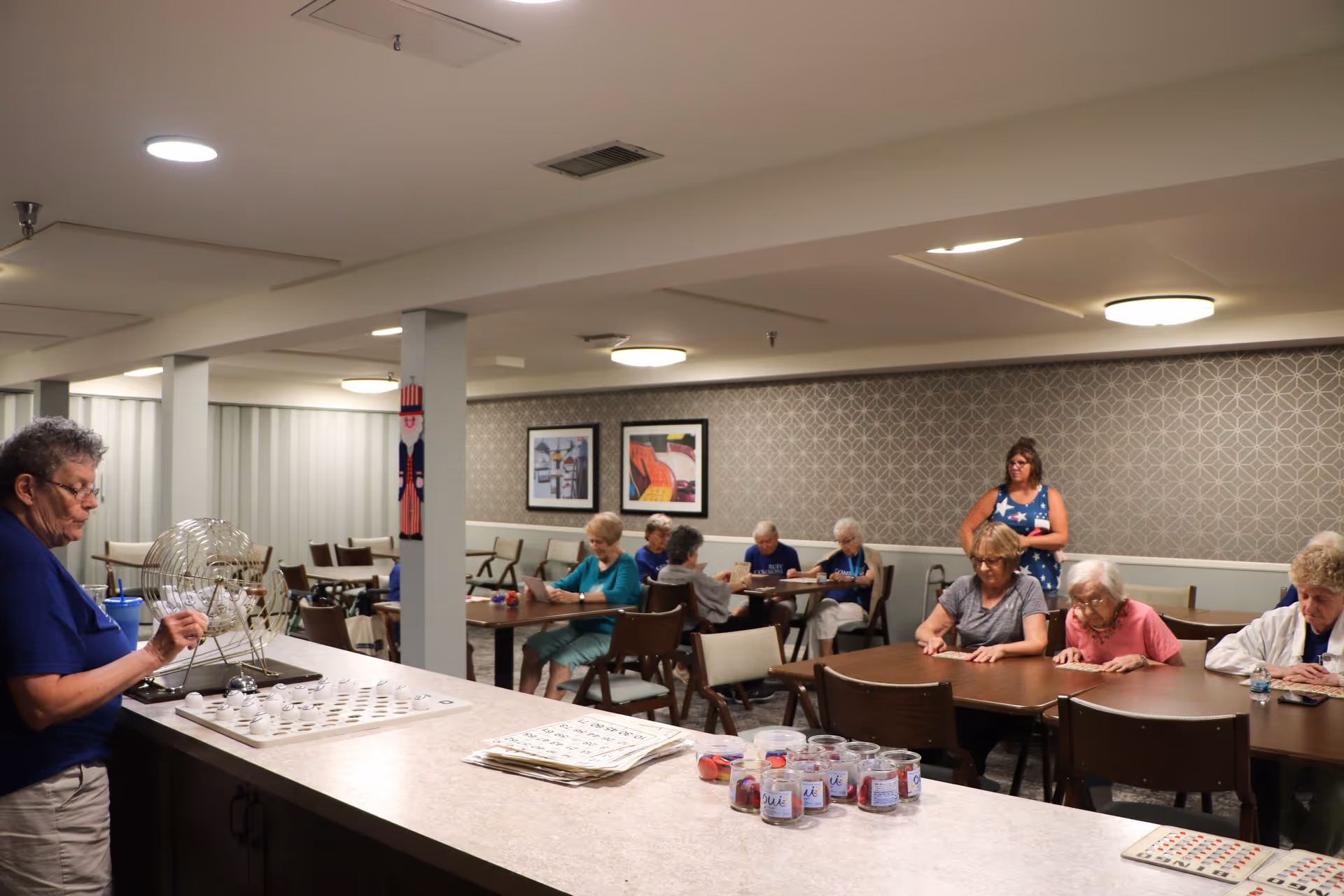 A group of elderly people seated at tables in a common room playing bingo. One person is standing behind the tables, and another person is operating a bingo cage at a counter in the foreground. The room has patterned wallpaper, framed artwork, and overhead lighting.