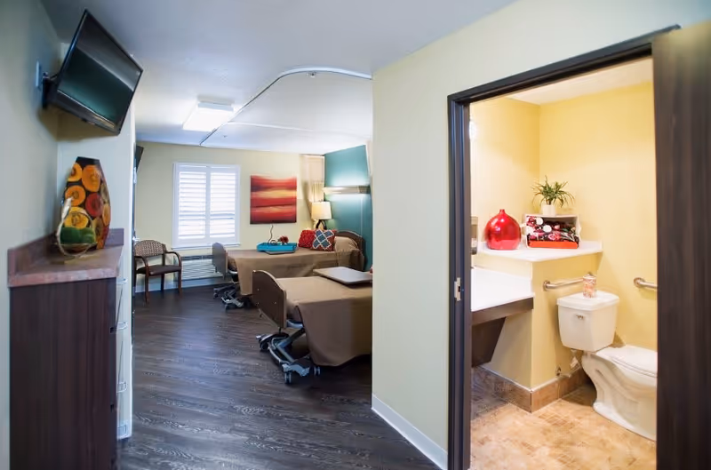 Interior view of a senior living resident room with two beds, seating, wall art, and an open bathroom to the right.