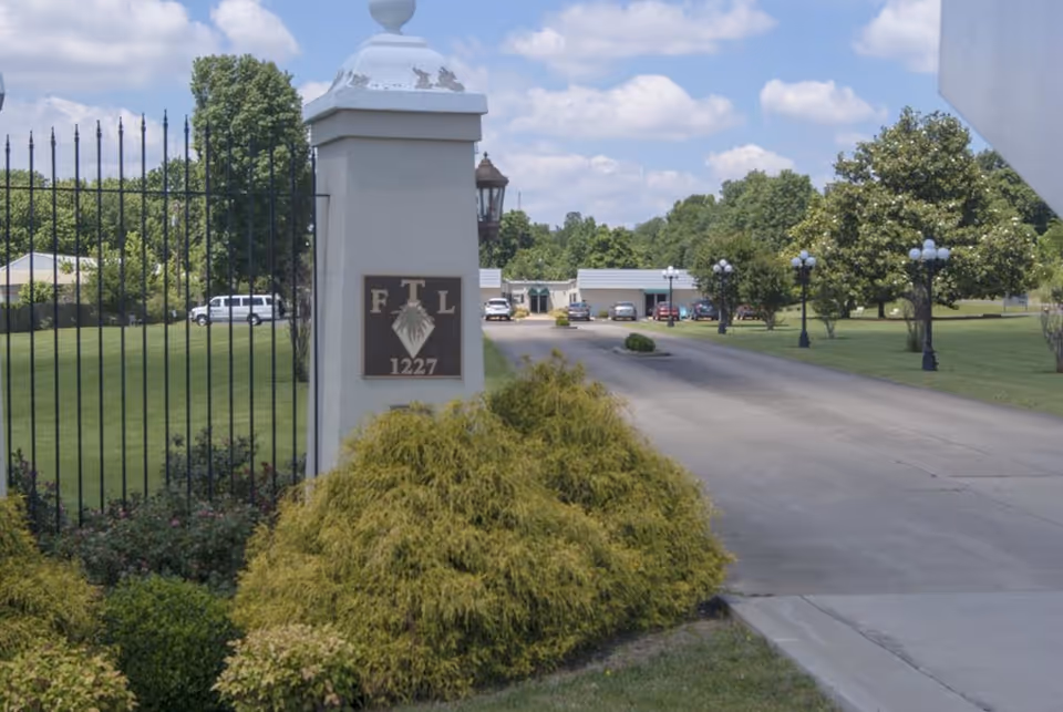 Entrance driveway with a gated pillar displaying a plaque reading 'FTL 1227', landscaped shrubs, lamp posts, and buildings in the background.