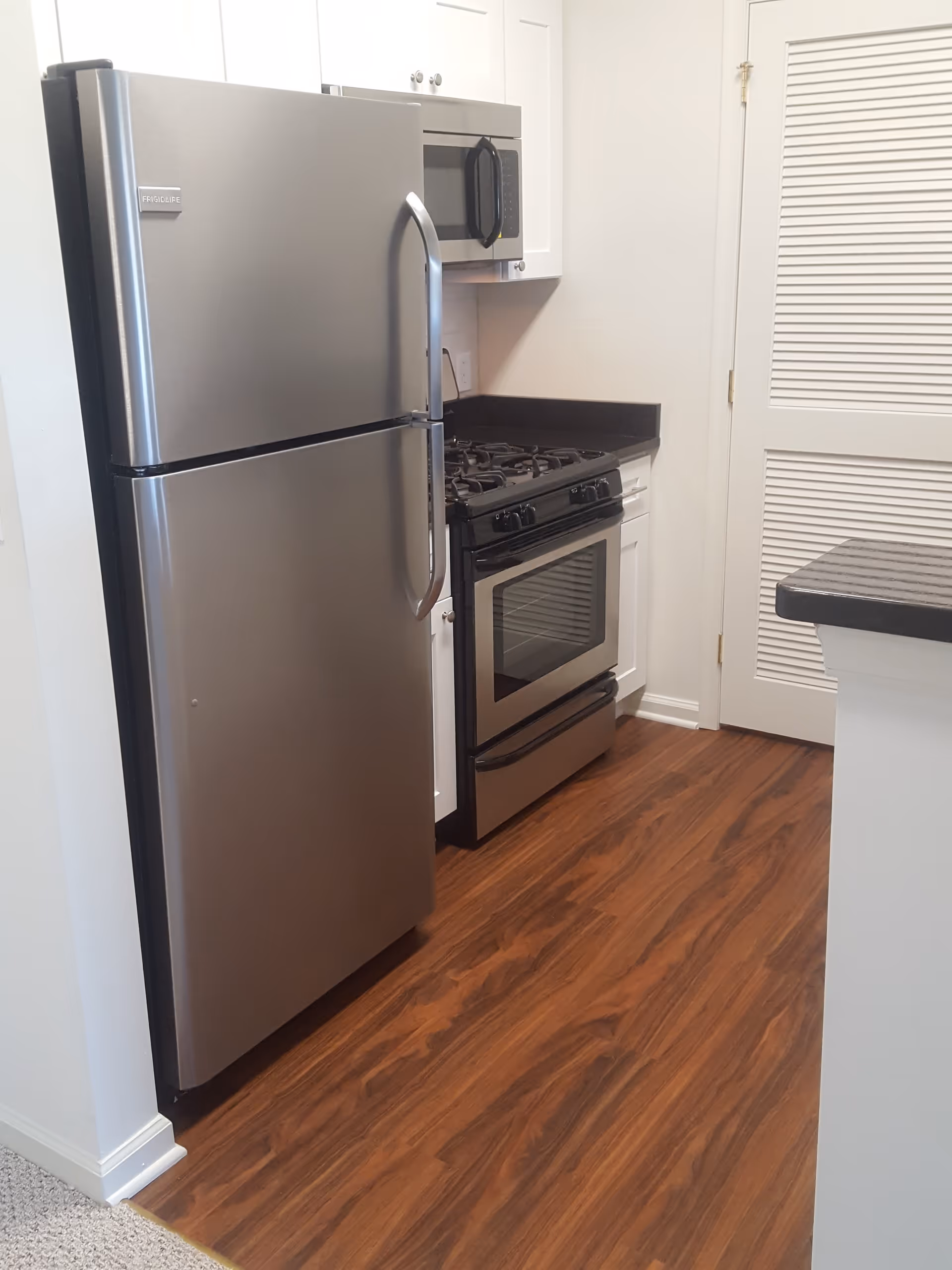A modern kitchen area featuring a stainless steel refrigerator, a gas stove with an oven, a microwave mounted above the stove, white cabinetry, and wood-patterned flooring.