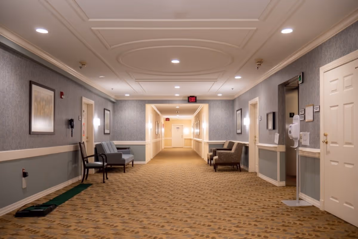 A well-lit hallway in a senior living facility with patterned carpet, gray and white walls, framed artwork, and several chairs placed along the walls. There is an elevator on the right side with a hand sanitizer dispenser nearby, and an exit sign is visible at the far end of the corridor.