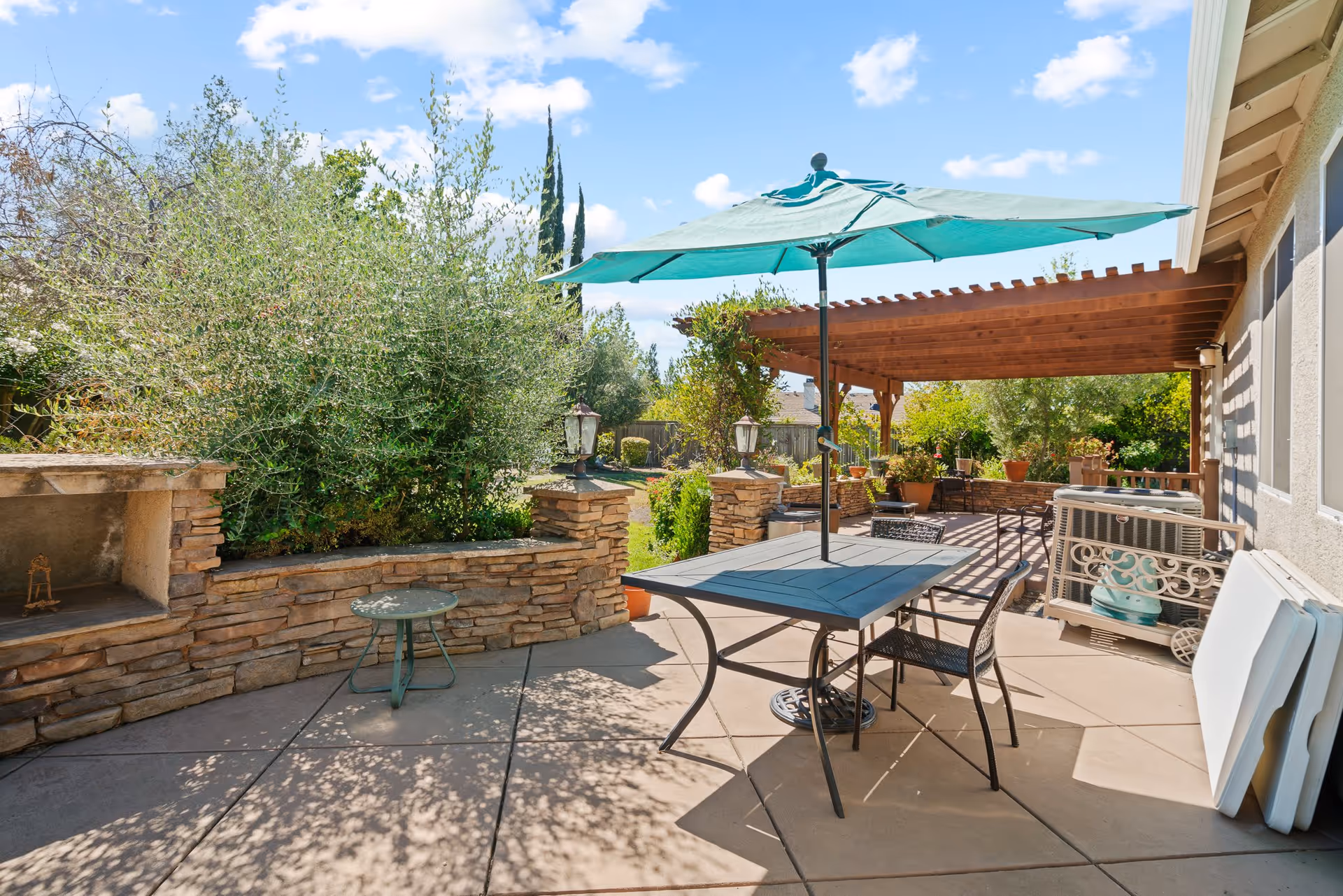 Outdoor patio area with a stone wall, a table with a teal umbrella, two chairs, a small round side table, and a wooden pergola with potted plants underneath. The patio is adjacent to a building with windows and an air conditioning unit. The sky is blue with some clouds.