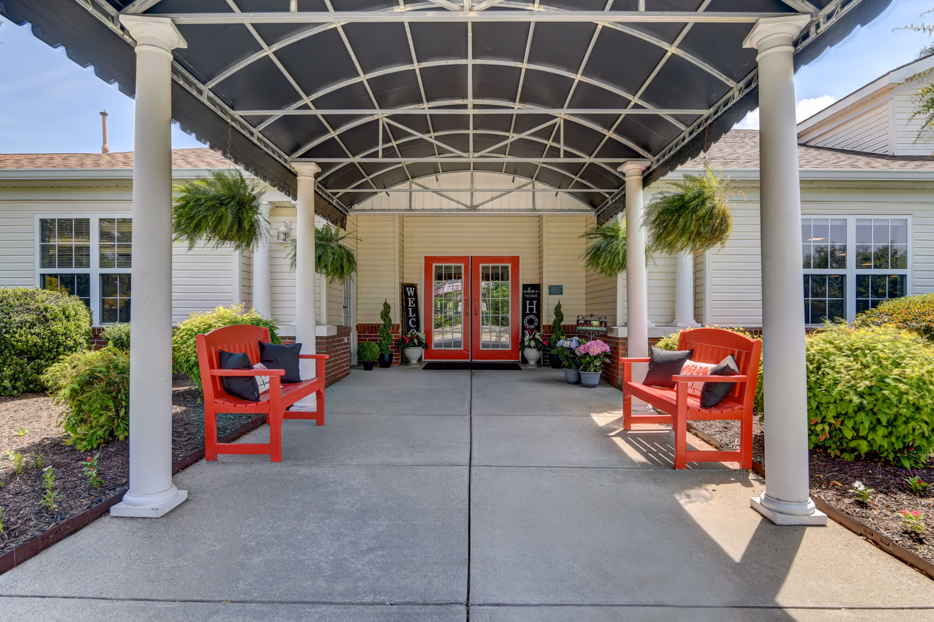 Covered entrance with columns, red benches, hanging plants, and red double doors leading into a senior living facility.