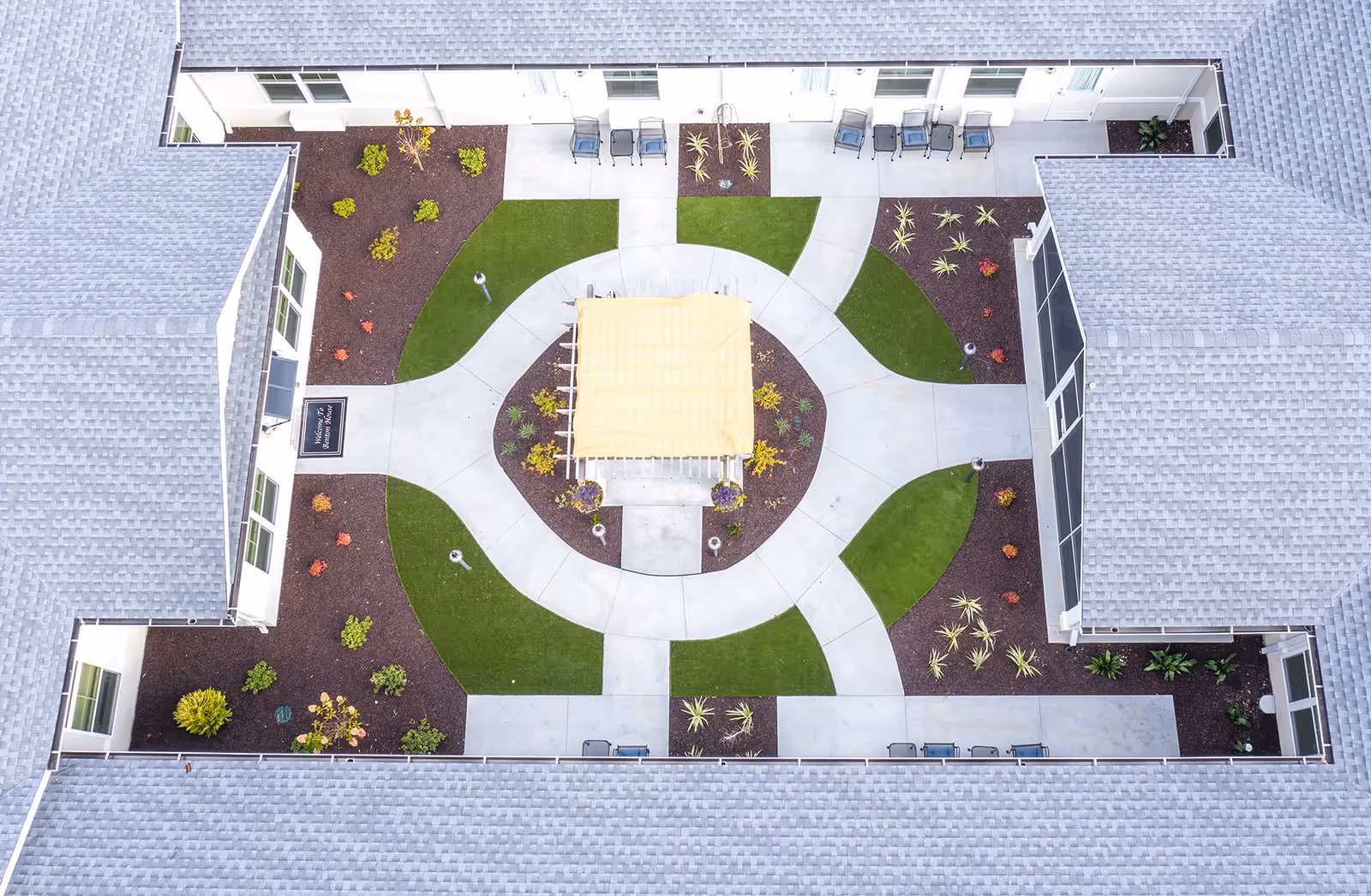 Aerial view of a courtyard in a senior living facility with a central pergola covered by a yellow canopy, surrounded by concrete walkways, green grass patches, and landscaped garden beds with various plants. The courtyard is enclosed by a building with gray shingled roofs and white walls, featuring multiple windows and outdoor seating areas with chairs.