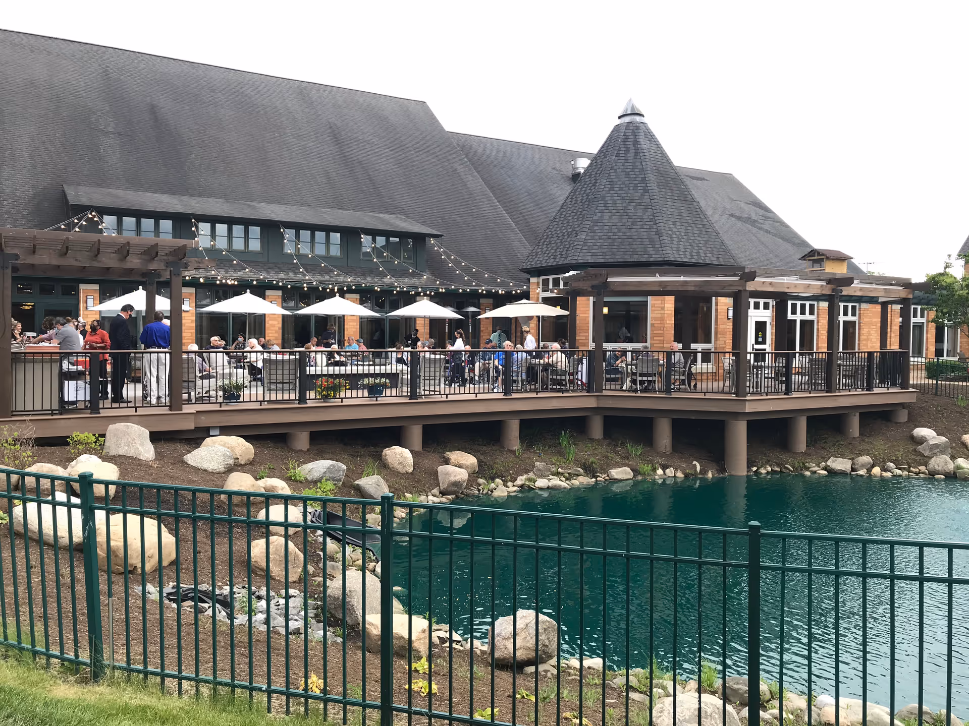 Outdoor patio area of The Summit at Brighton Independent Living with people sitting under white umbrellas at tables on a wooden deck next to a pond with rocks and a black metal fence in the foreground. The building has a steep roof and large windows, with string lights hanging above the patio.