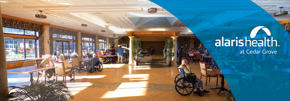 A spacious, sunlit common area in a senior living facility with large windows along one side. Several elderly residents are seated at tables and in wheelchairs, engaging in conversation or relaxing. The room features wooden flooring, decorative columns, and potted plants. The right side of the image has a blue overlay with the text 'alaris health at Cedar Grove'.