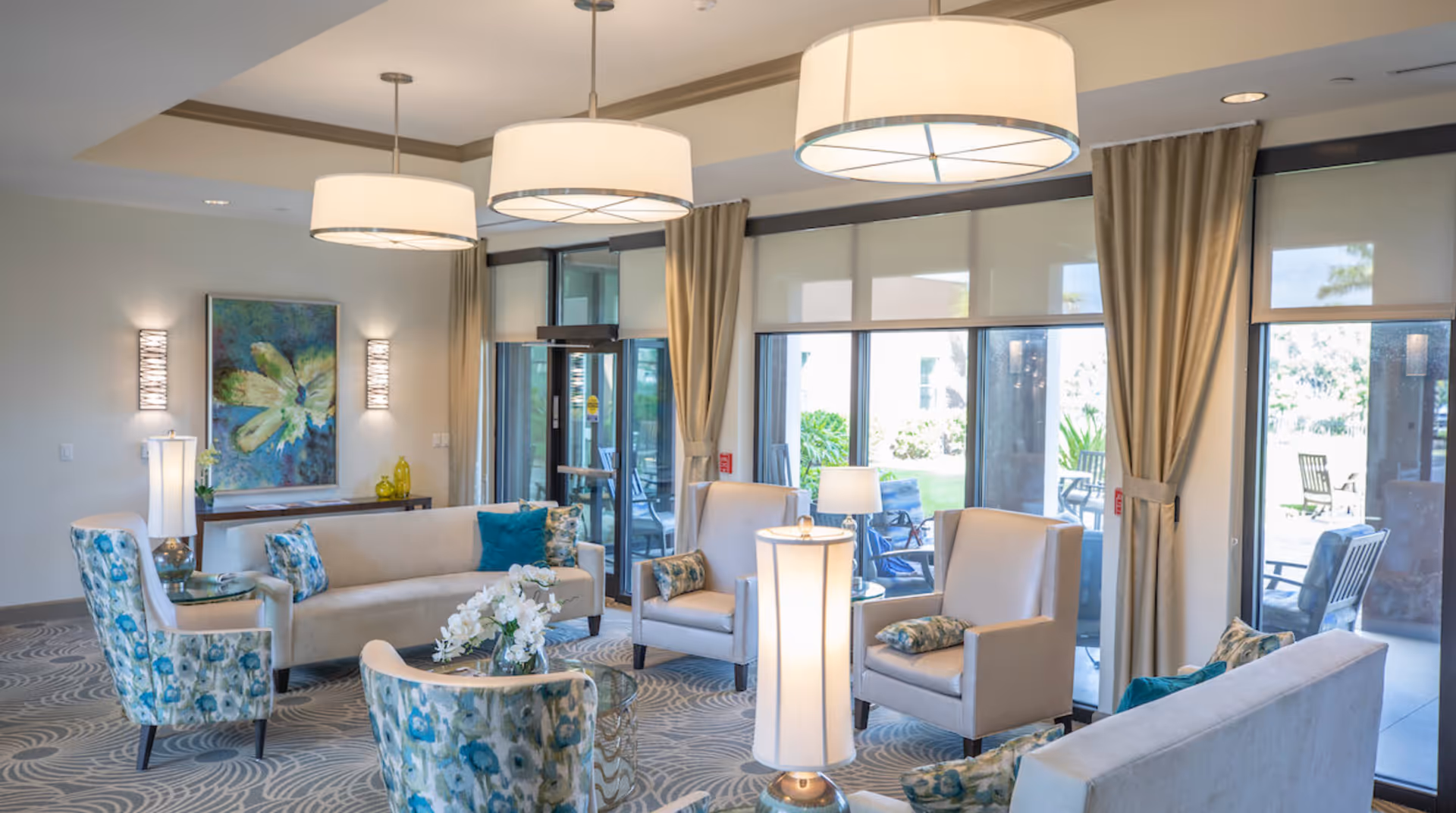 Bright, modern senior living lounge with sofas, patterned armchairs, pendant lights and large windows overlooking outdoor seating.