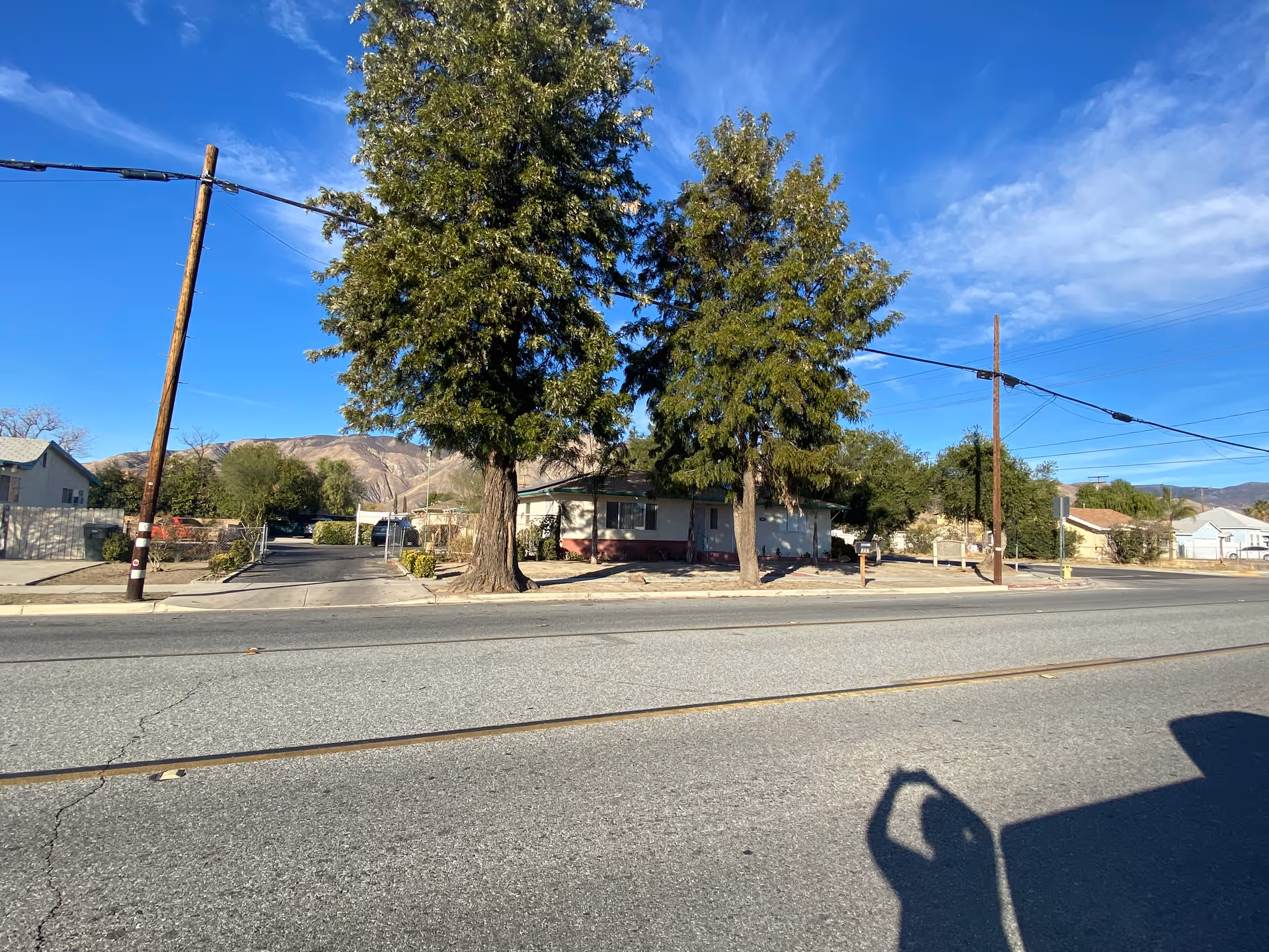Street view of a residential area with a single-story house partially obscured by two large trees. There are utility poles and wires along the street, with mountains visible in the background under a blue sky with some clouds.