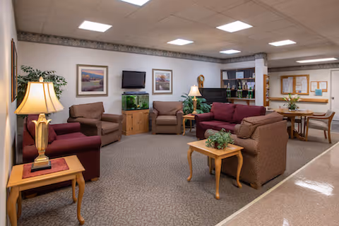 A cozy common area in a senior living facility with several upholstered chairs and sofas arranged around wooden side tables. There are two table lamps, framed artwork on the walls, a fish tank on a wooden stand, and a bulletin board in the background. The room has a carpeted floor and fluorescent ceiling lights.
