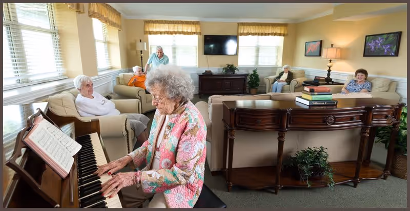 A group of elderly women in a bright living room at a senior living facility. One woman is playing the piano while others sit comfortably on sofas and chairs, listening and enjoying the music. The room has large windows with blinds, a flat-screen TV mounted on the wall, and decorative plants and artwork.