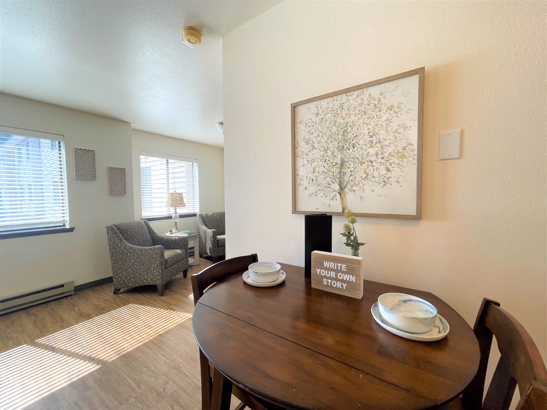 A bright living area with two patterned armchairs near two windows with blinds. A small round wooden dining table with two place settings and a decorative sign that reads 'WRITE YOUR OWN STORY' is in the foreground. A framed artwork of a tree hangs on the wall above the table.