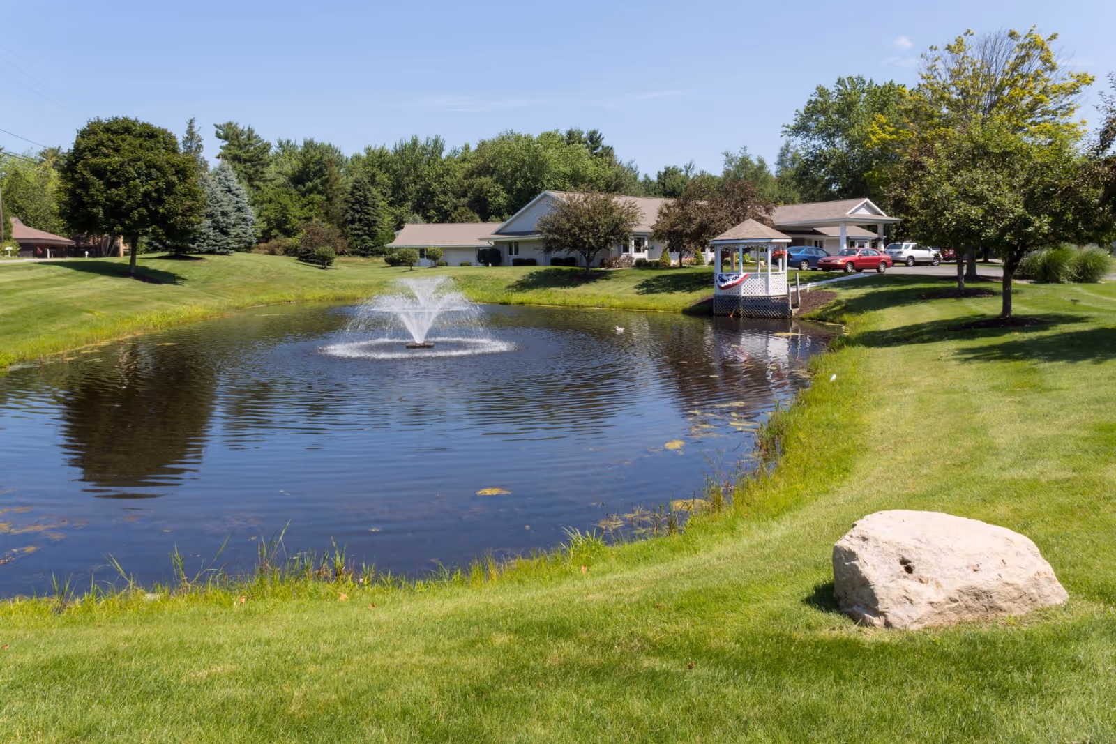 Lush lawn surrounds a pond with fountain, gazebo, and a building at American House Spring Lake.