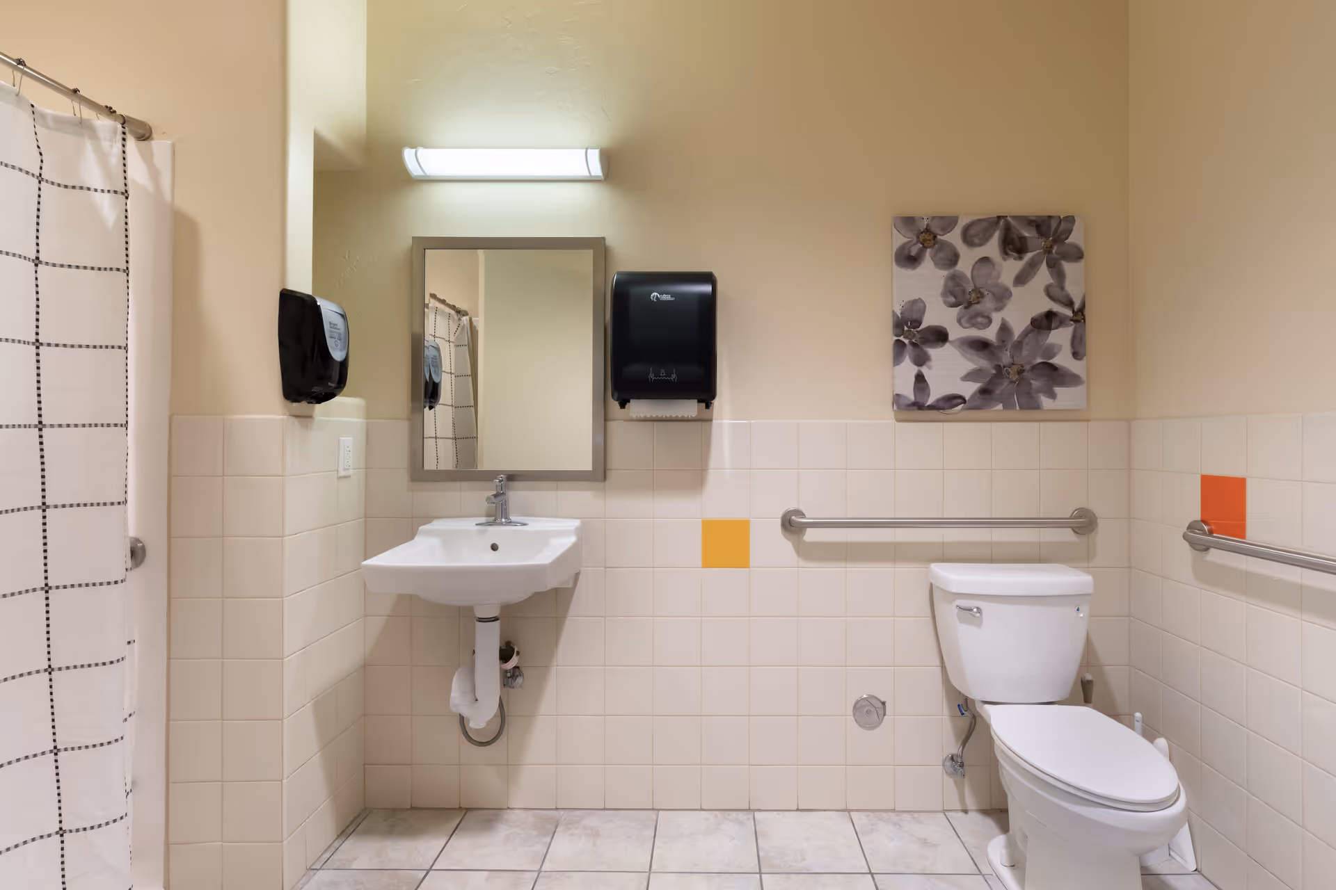 A clean and accessible bathroom with beige tiled walls and floor. The room features a white toilet with grab bars on both sides, a white wall-mounted sink with a mirror above it, a soap dispenser, and a paper towel dispenser. There is a shower area with a white curtain featuring a black grid pattern. A floral artwork hangs on the wall above the toilet.