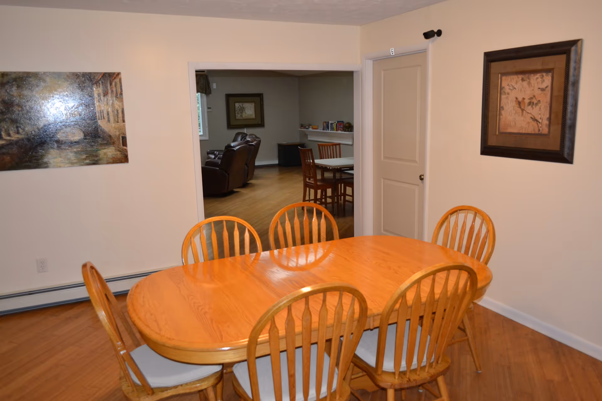 A dining room with a wooden oval table surrounded by six wooden chairs with light gray cushions. The room has light-colored walls with two framed paintings hanging on them. Through an open doorway, a living room area with brown leather recliners and another table with chairs is visible. There is a closed door labeled with the number 6 on the right side of the dining room.