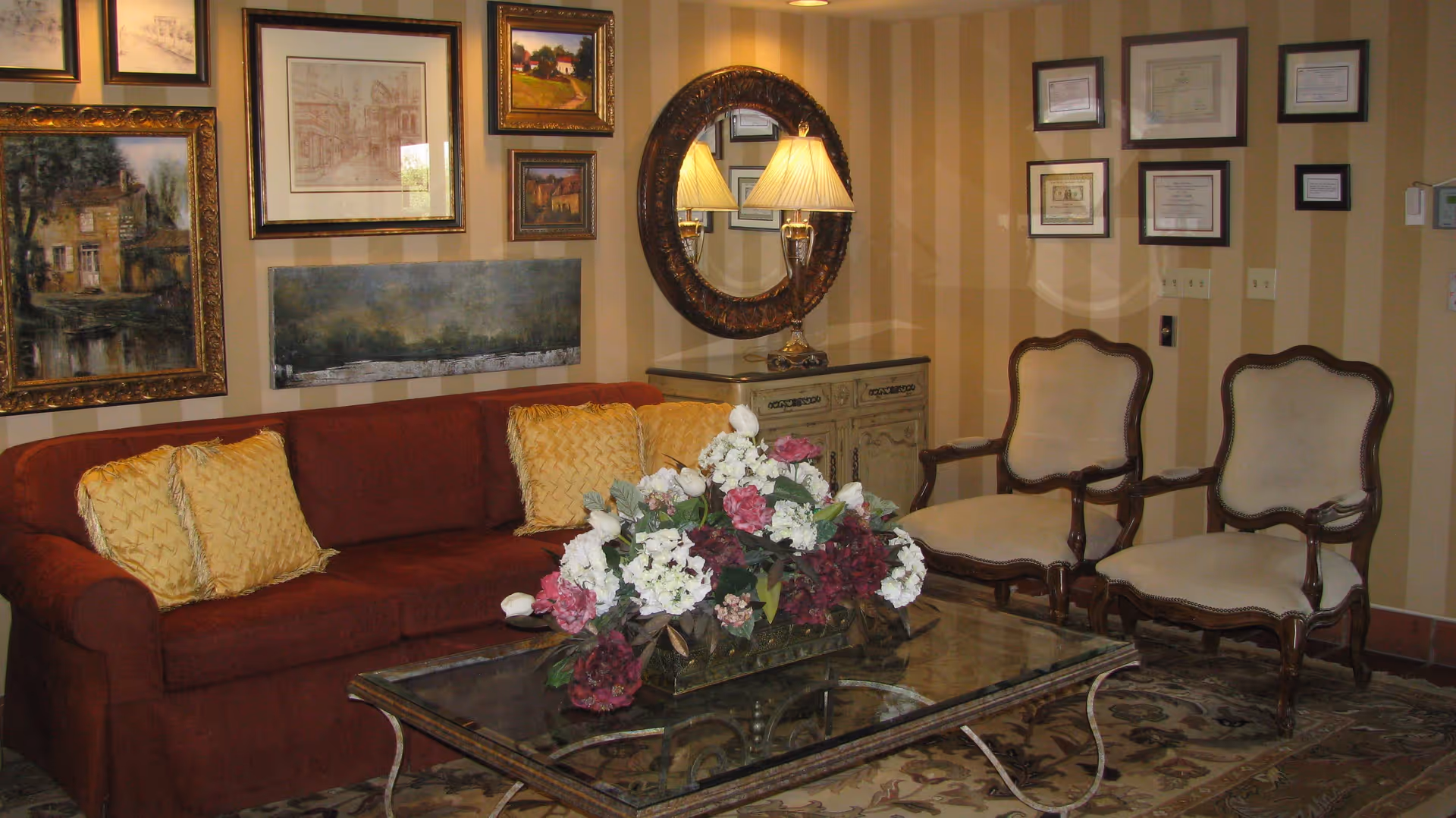 Cozy seating area with a red sofa, two armchairs, a glass coffee table topped with a floral arrangement, and framed artwork on striped walls.