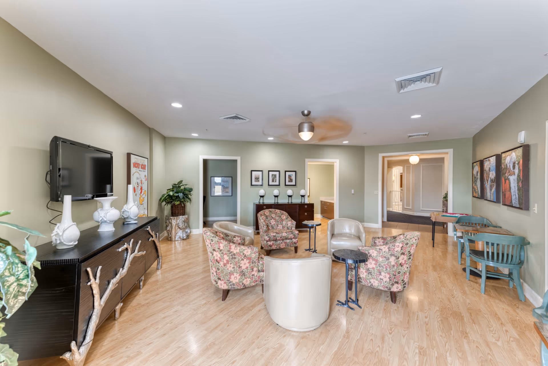 A spacious and well-lit common area with light wood flooring and light green walls. The room features a circular seating arrangement with floral-patterned and beige chairs around small black tables. On the left wall, there is a black cabinet with decorative white vases and a mounted flat-screen TV above it. The back wall has three framed pictures and a sideboard with four spherical lamps. To the right, there are two small tables with green chairs and colorful artwork on the wall. The ceiling has recessed lighting and a ceiling fan with a light.